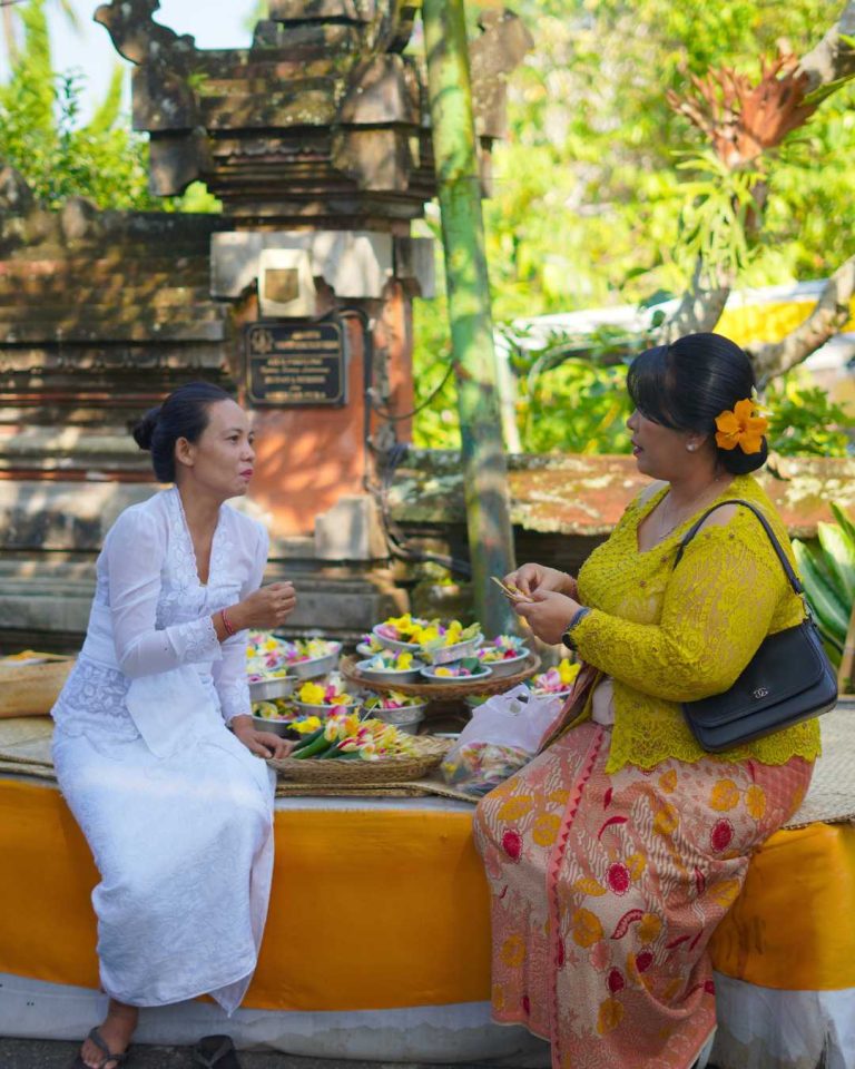 Two Balinese women preparing offerings and sharing a conversation during a temple ceremony in Bali.